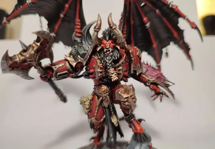 Groźny chaos daemon prince z czerwono-złotą zbroją, czarnymi skrzydłami i toporem.