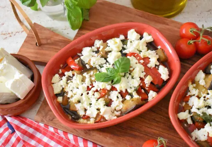 Pyszna pieczona feta z warzywami, podana w glinianym naczyniu, z dodatkiem świeżej bazylii i pomidorków.