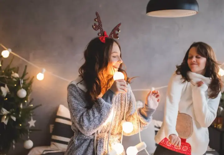 Due ragazze cantano canzoni di Natale in inglese con testo, tenendo una lampadina come microfono, circondate da luci natalizie e un albero addobbato.
