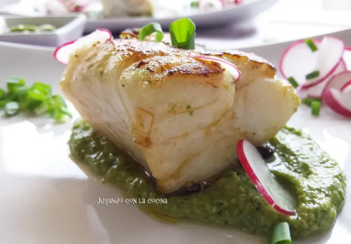 Un trozo de pescado blanco y tierno reposa sobre una cama de vibrante mojo verde, adornado con rodajas de rábano y cebolleta.