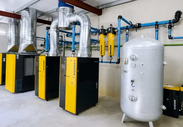 Instalación industrial con compresores y secadores de aire comprimido, tuberías azules y un gran tanque de almacenamiento plateado.