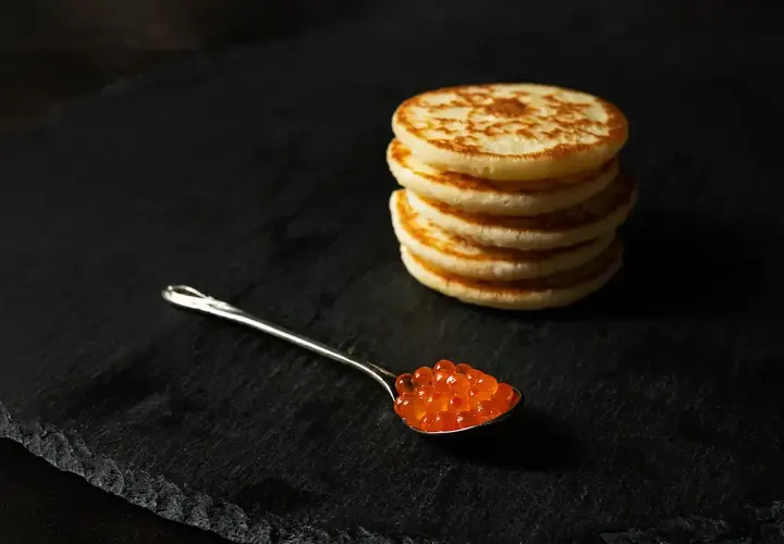 Une cuillère de caviar rouge sur une assiette noire, à côté d'une pile de blinis dorés. Parfait pour une recette blinis gourmande.