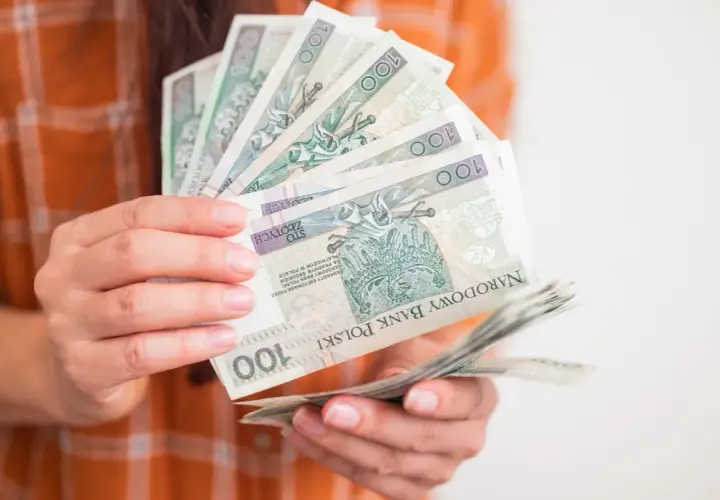 Dłonie trzymają wachlarz banknotów 100 zł. Taka kwota to już całkiem spora kuroniówka na drobne wydatki.