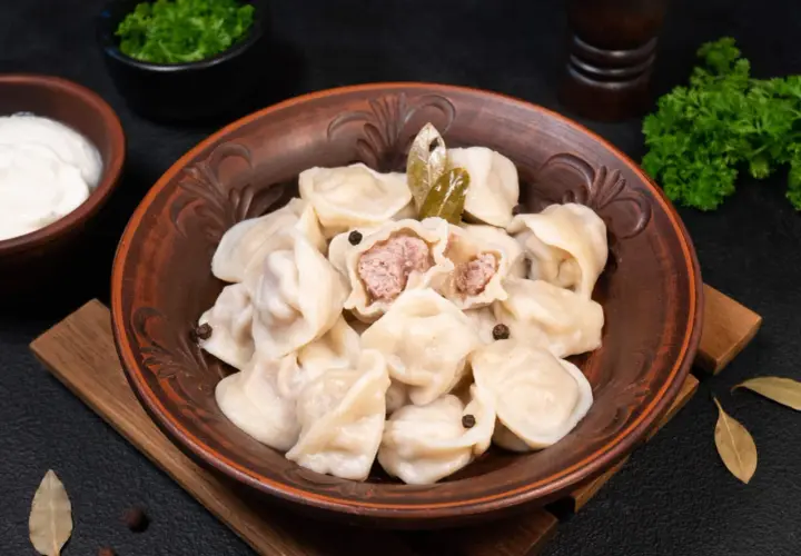 Pielmieni co to? Pyszne, domowe pierożki z mięsem, podane w glinianej misie z liśćmi laurowymi i pieprzem. Obok śmietana i natka pietruszki.