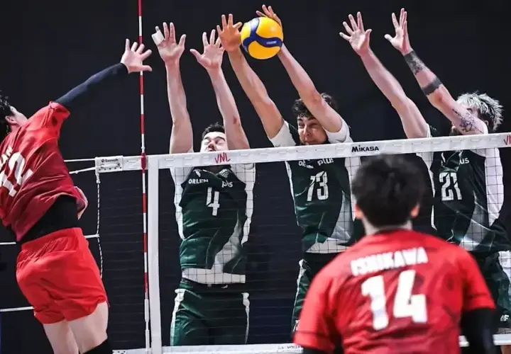 Zacięta akcja siatkarska: polska Japonia w akcji. Zawodnik w czerwonej koszulce atakuje, blokujący w zielonych strojach próbują zatrzymać piłkę.