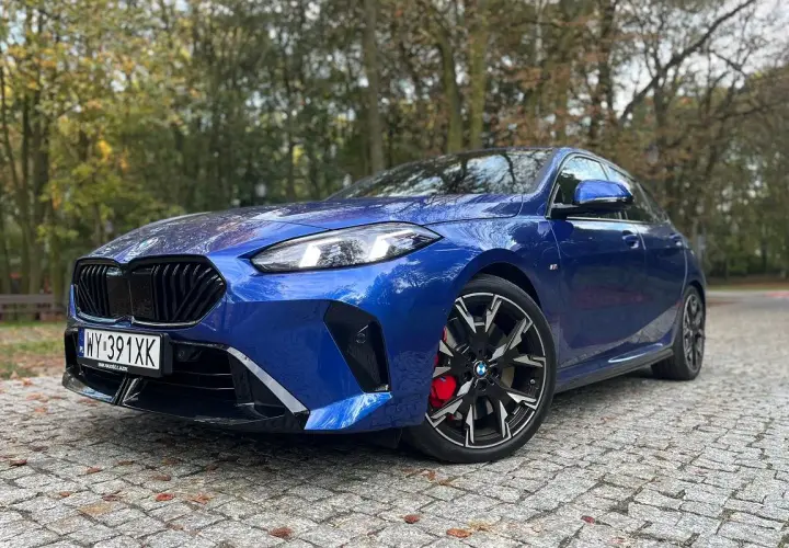 Błękitne BMW 235i z czarnymi felgami i czerwonymi zaciskami hamulcowymi zaparkowane na bruku wśród jesiennych drzew.
