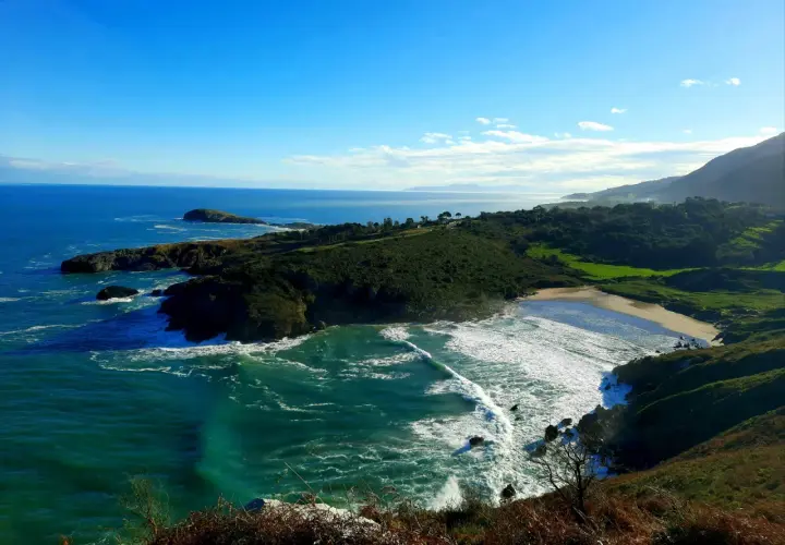 Vista panorámica de la playa de Sonabia, con olas rompiendo en la costa rocosa y un mar azul intenso bajo un cielo despejado.