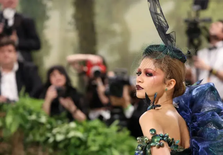 Una modelo con un elaborado atuendo de **Met Gala theme**, con velo y maquillaje dramático, posa rodeada de fotógrafos y vegetación.