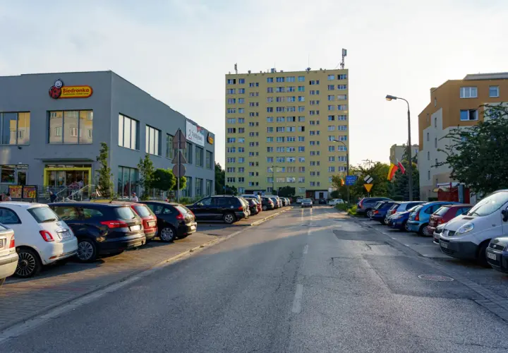 Uliczka z zaparkowanymi samochodami obok sklepu Biedronka, gdzie logo przypomina uśmiechniętego ursusa niedźwiadka. W tle wysoki, żółty blok mieszkalny.
