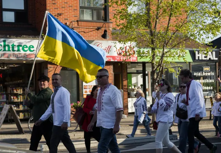 Ludzie w tradycyjnych strojach niosą flagę Ukrainy. W tle sklepy, symbolizujące życie codzienne, które wciąż trwa, mimo trudności zagrażających liczba ludności Ukrainy.