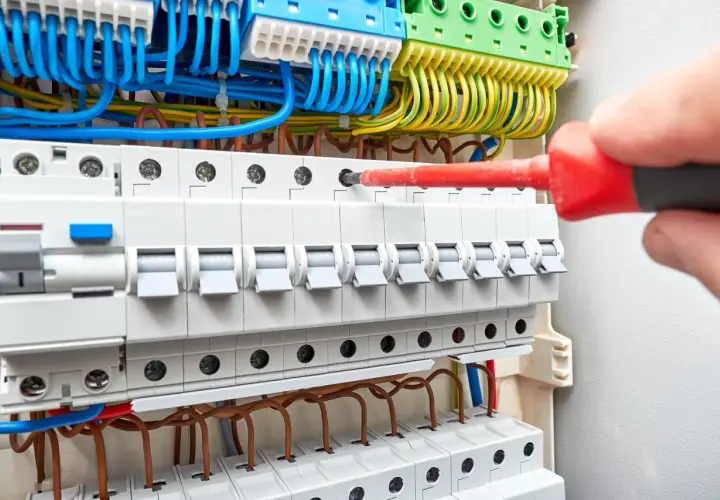 Ręka z czerwonym śrubokrętem przykręca przewody w skrzynce elektrycznej. Trwa wymiana instalacji elektrycznej.