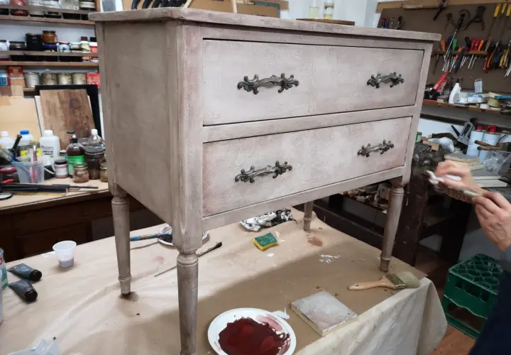 Un mobile antico in fase di restauro, con cassetti e pomelli decorati. Attrezzi e materiali da lavoro circondano il pezzo.