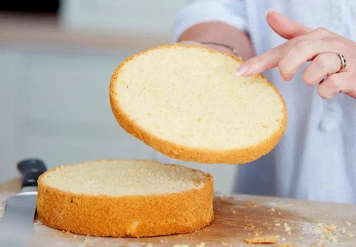Perfekter tortenboden rezept: Eine Hand hebt eine goldbraune Kuchenschicht über eine zweite, die auf einem Holzbrett liegt.