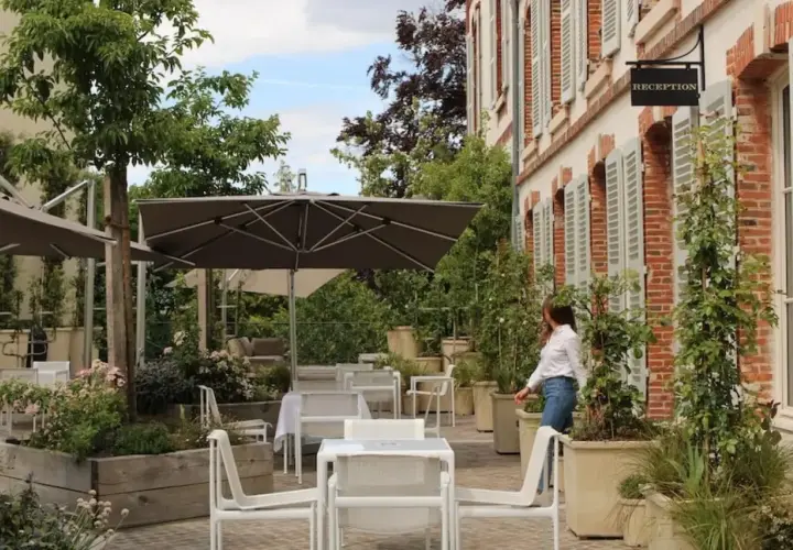 Terrasse verdoyante du 25bis by Leclerc Briant, avec tables, chaises et parasols. Une femme se dirige vers la réception.