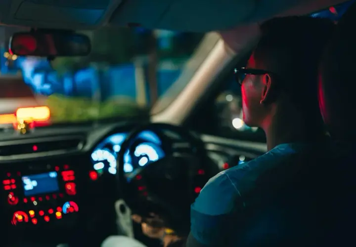 Un hombre con gafas conduce de noche, con el tablero iluminado en azul y rojo.