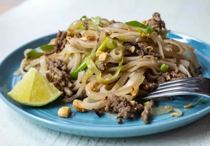 Pyszny pad thai przepis na talerzu: makaron ryżowy z mielonym mięsem, cebulą, orzeszkami ziemnymi i limonką.