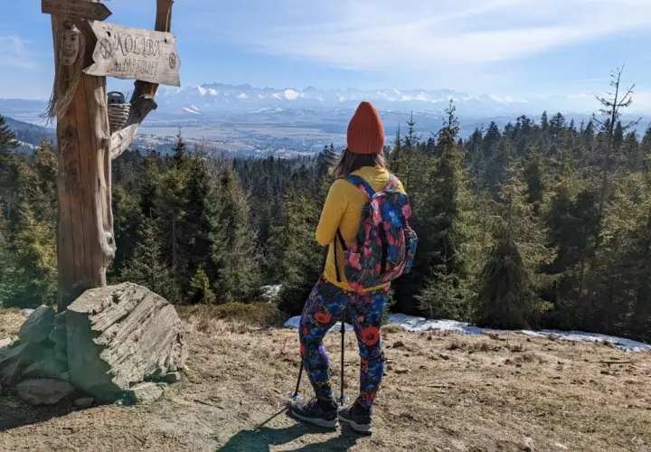 Widok na Tatry z drewnianego słupa z napisem "Koliba na Łapsowej Polanie". Turystka w kolorowych legginsach podziwia panoramę.