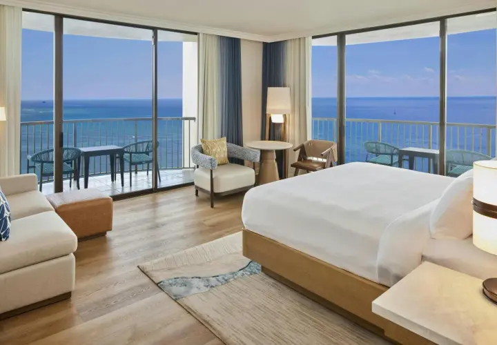 Pokój hotelowy z widokiem na ocean, idealny na wakacje hawaje all inclusive. Wygodne łóżko, sofa i balkon z stolikiem.