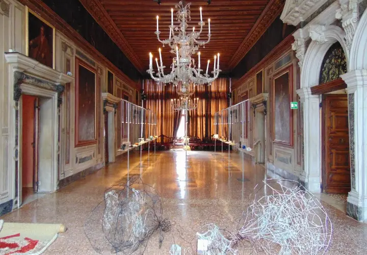 Intérieur du Palazzo Mocenigo, un long couloir orné de tableaux, avec un lustre imposant et des sculptures modernes au sol.