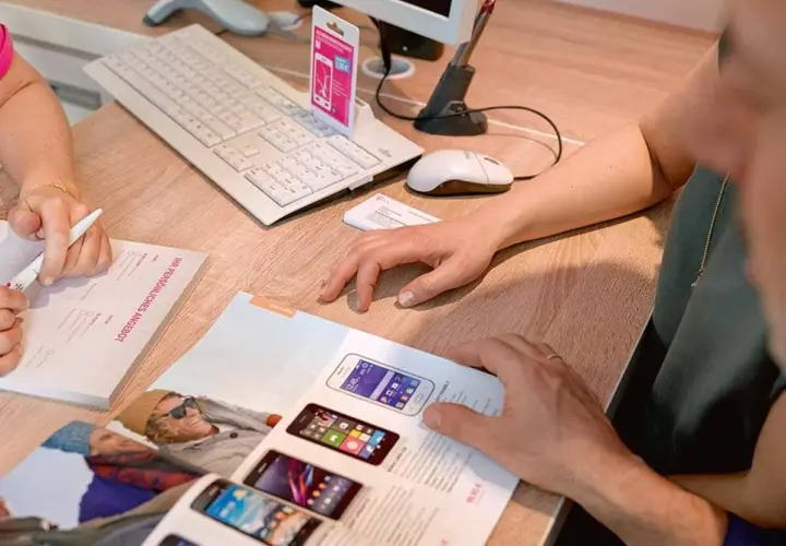 Klienci przeglądają ofertę smartfonów T-Mobile. Na biurku klawiatura, myszka i dokumenty.