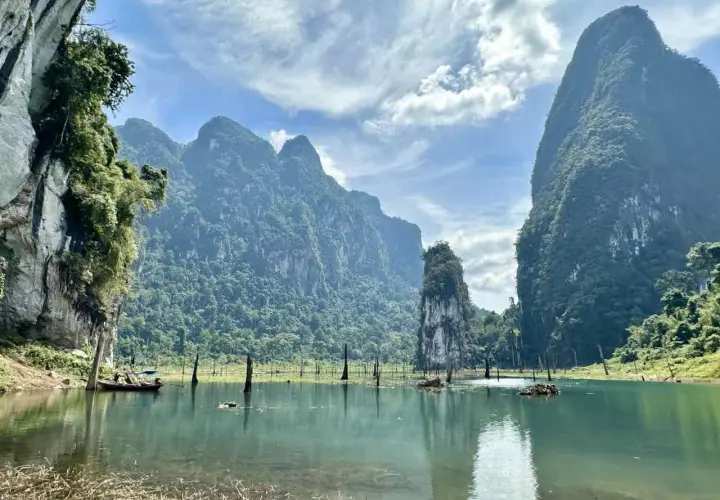 Khao Sok Nationalpark: Majestätische Kalksteinfelsen ragen aus einem ruhigen See empor, umgeben von üppigem Dschungel. Ein Boot gleitet über das Wasser.