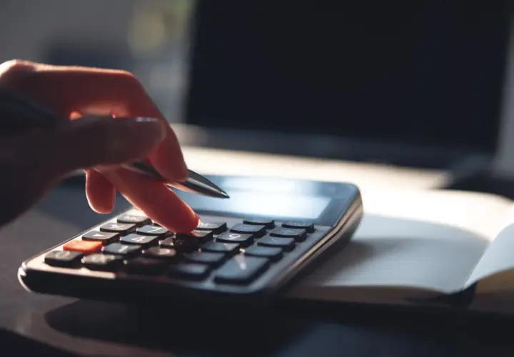Dłoń z długopisem naciska klawisze kalkulatora, licząc, ile zarabia 1% najbogatszych Polaków. Obok leży otwarty notatnik i laptop.