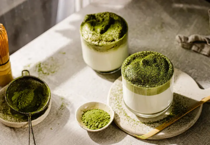 Zwei Gläser cremiger Matcha-Latte, bestreut mit feinem Pulver. Die Zubereitung mit Chasen und Sieb deutet auf die belebende matcha wirkung hin.