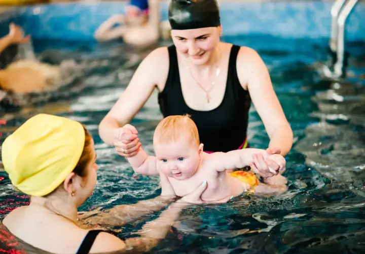 Radość na twarzy dziecka podczas zajęć na basenie dla niemowląt. Opiekunka trzyma malucha, który z zaciekawieniem patrzy przed siebie.