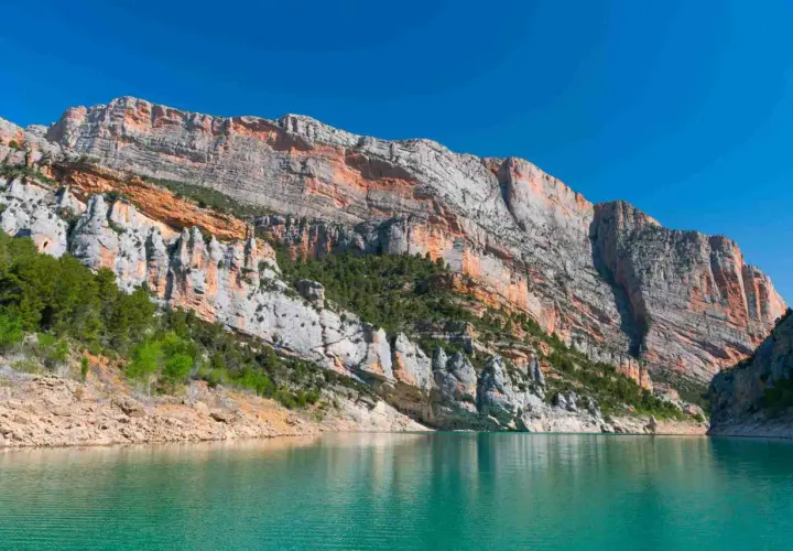 Majestuosas paredes rocosas de la sierra del Montsec se reflejan en las aguas turquesas de un embalse.