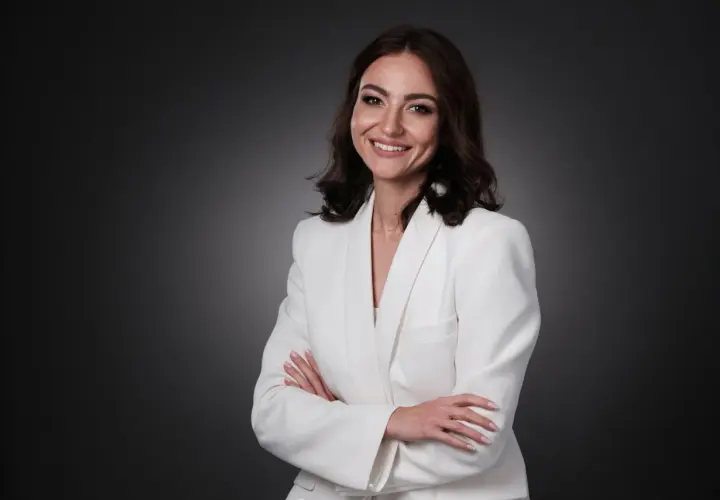 Uśmiechnięta kobieta w białej marynarce, z rękami skrzyżowanymi na piersi. Idealna sesja biznesowa pozy.