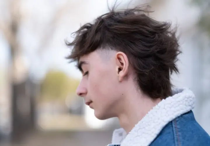 Junger Mann mit modernen Mullets, der nachdenklich zur Seite blickt. Sein Haar ist dunkelbraun und zerzaust, die Seiten sind kurz geschoren. Er trägt eine Jeansjacke mit Teddyfellkragen.