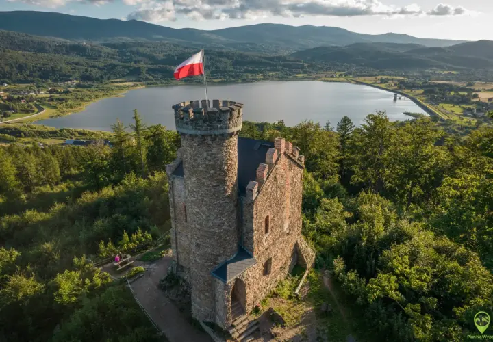 Kamienny zamek henryka z polską flagą na wieży, otoczony zielenią, z jeziorem i górami w tle.