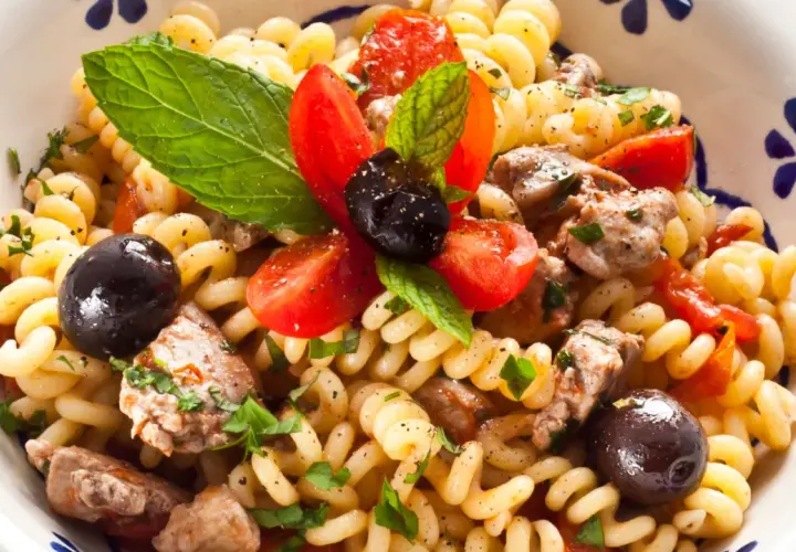 Pasta con pesce spada, pomodorini, olive nere e menta fresca in una ciotola decorata.