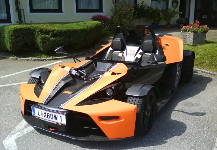 Pomarańczowy KTM X-Bow z czarnymi akcentami i tablicą rejestracyjną "XBOW 1" stoi na parkingu przed budynkiem z zielonym żywopłotem.