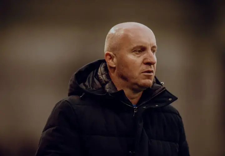 Nowy trener Widzewa, łysy mężczyzna w czarnej kurtce, patrzy w bok z zamyśloną miną.