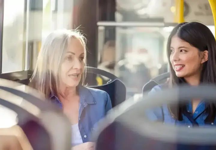 Dwie kobiety, starsza i młodsza, siedzą obok siebie w autobusie. Starsza, z siwymi włosami, opowiada coś młodszej, która słucha z uśmiechem. To miła rozmowa z nieznajomym.