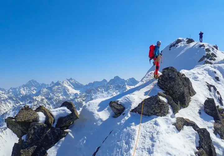 Wspinaczka zimą na Orlą Perć. Dwóch alpinistów na ośnieżonym szczycie, jeden z nich zabezpiecza się liną.