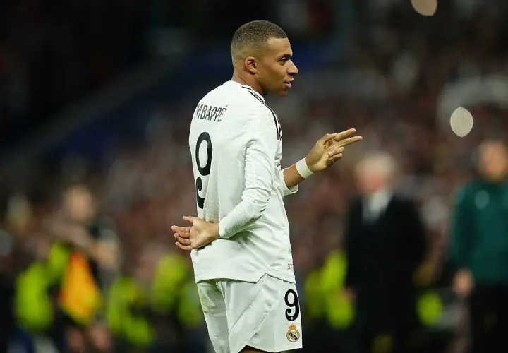 Mbappe, w białej koszulce z numerem 9, gestykuluje na boisku. Jego wzrost robi wrażenie, a kibice śledzą każdy jego ruch.