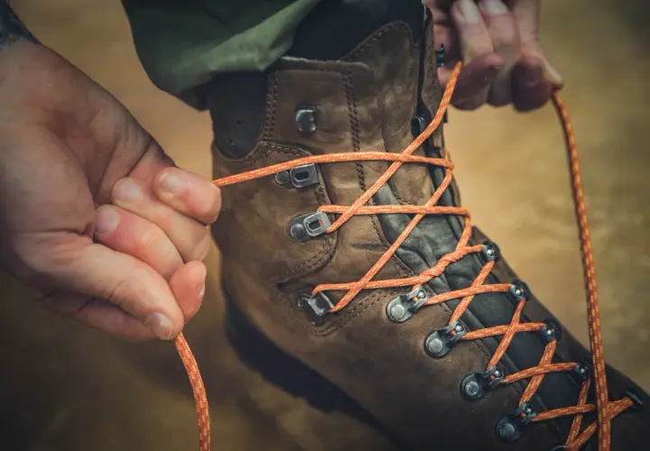 Close-up of hands tying orange laces through metal hooks on a brown hiking boot, demonstrating how to tie boots with hooks for a secure fit.
