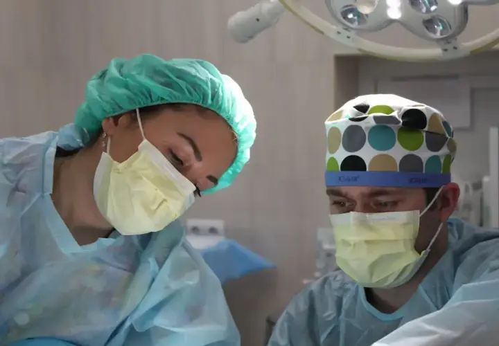 Pielęgniarka i chirurg w maseczkach i czepkach pracują pod lampą operacyjną.