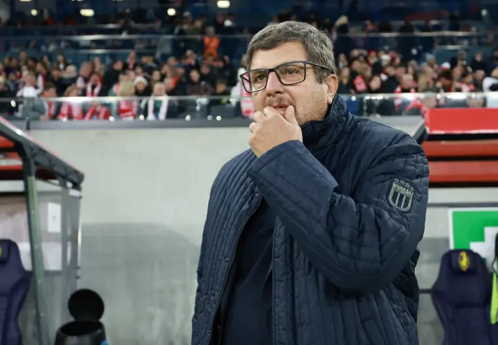 Włoski trener w okularach, zamyślony, w granatowej kurtce z naszywką "ITALIA", stoi na tle trybun stadionu.