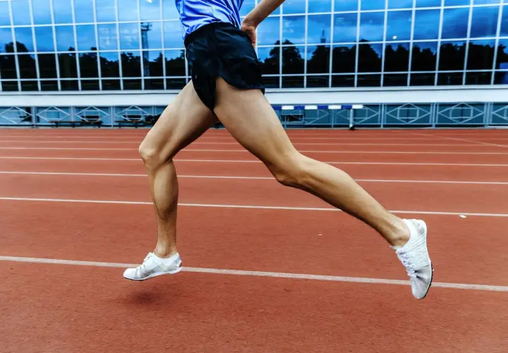 Sylwetka biegacza w ruchu na bieżni stadionu. Mięśnie nóg naprężone, białe buty sportowe unoszą się nad czerwonym torem.