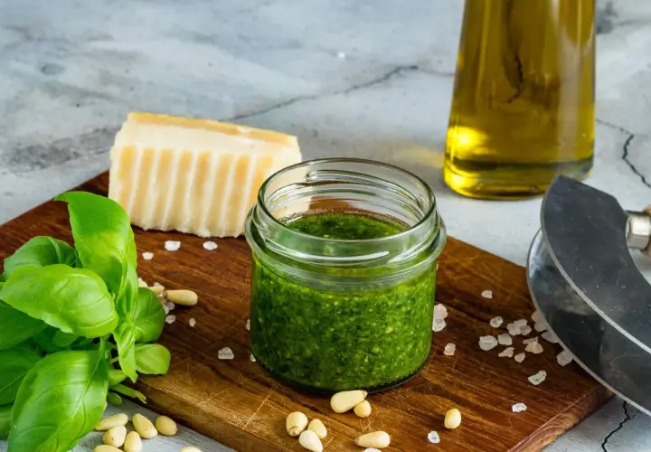 Słoiczek domowego pesto z bazylii, idealny sposób, jak zrobić pesto z bazylii na zimę. Obok świeża bazylia, orzeszki piniowe, parmezan i oliwa.