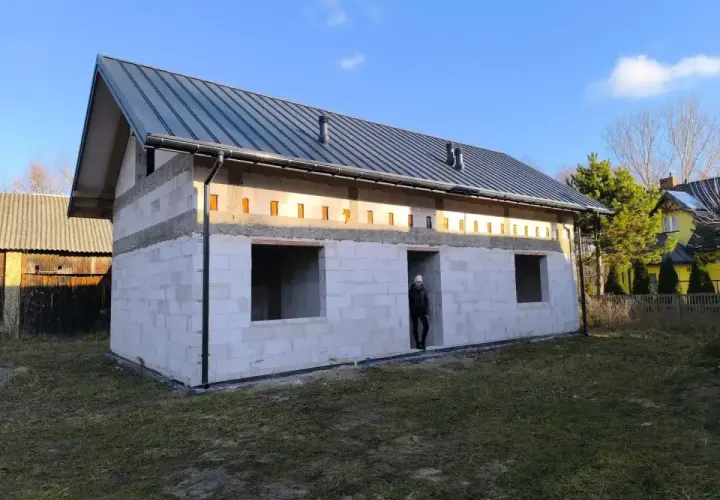 Budowa domu bez pozwolenia w toku. Białe bloczki, szary dach, puste okna. Ktoś stoi w drzwiach.