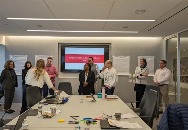 A team coach leads a meeting in a conference room, with a screen displaying "What will make today successful for you?".