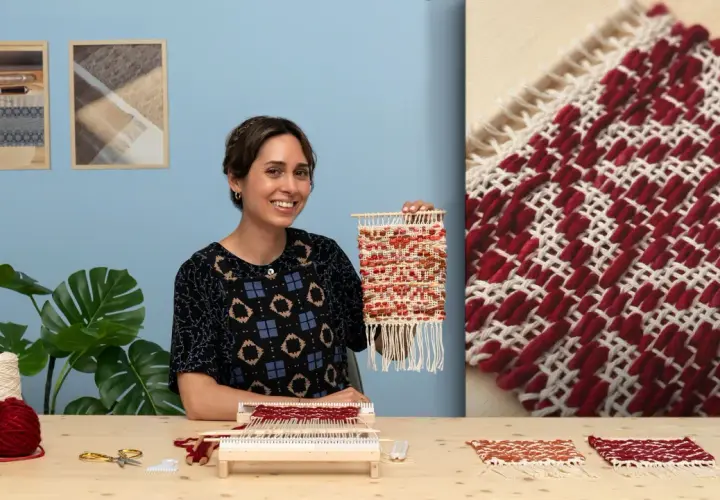 Artisanat textile : une femme souriante tient une petite tapisserie colorée. Sur la table, du fil, des ciseaux et un métier à tisser.