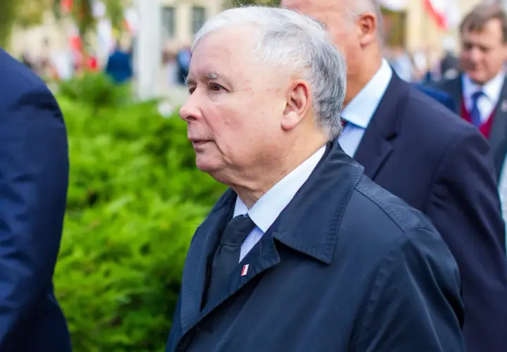 Jarosław Kaczyński, lider partii, w czarnym płaszczu i z przypinką z polską flagą, podczas oficjalnych uroczystości.