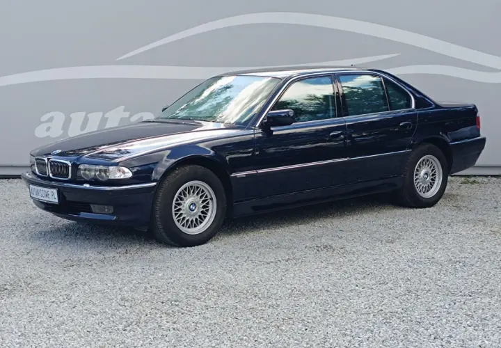 Ciemnoniebieskie BMW 7 E38 z lat 90. stoi na żwirowym podjeździe, z odbiciem chmur w szybach.