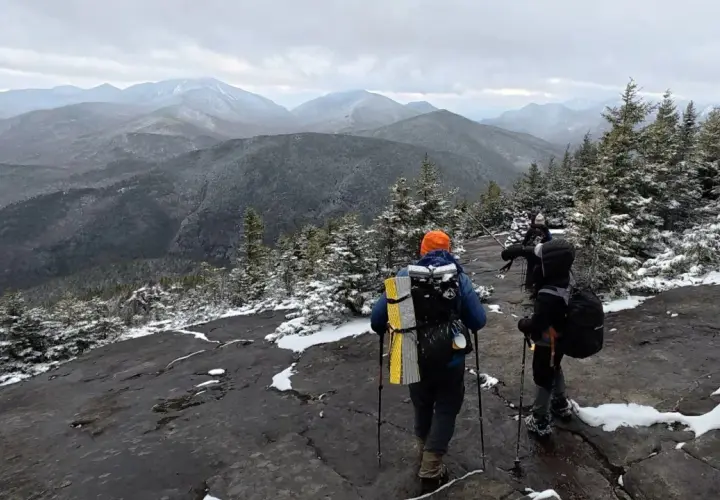 Hikers trek across a snowy mountain ridge, leaving no hiker trash behind. The vast, misty mountain range stretches into the distance under a cloudy sky.