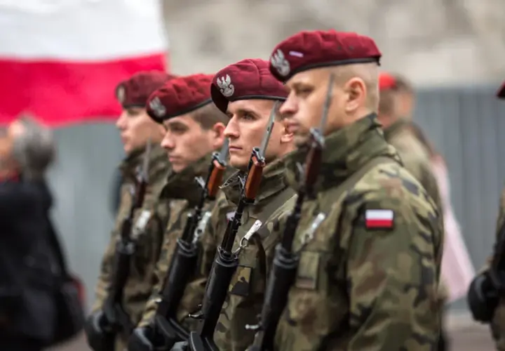 Żołnierze w bordowych beretach i mundurach moro, z karabinami, stoją w szeregu. W tle polska flaga. To dumne kategorie wojskowe.